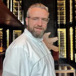 André Kaiser, Chef Culinaire im The Chedi Andermatt, in weißer Kochjacke vor Weinschränken und dunklem Interieur mit Holzskulptur
