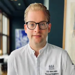 Thomas Anton Beiglboeck, Chef Culinaire im Les Trois Rois Basel, in weißer Kochjacke mit Brille vor stilvoll-modernem Innenraum