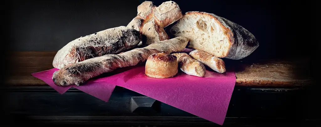 Verschiedene Brotsorten auf dunkler Holzplatte mit violettem Papier, darunter Flûtes, Focaccia und halbiertes Krustenbrot.