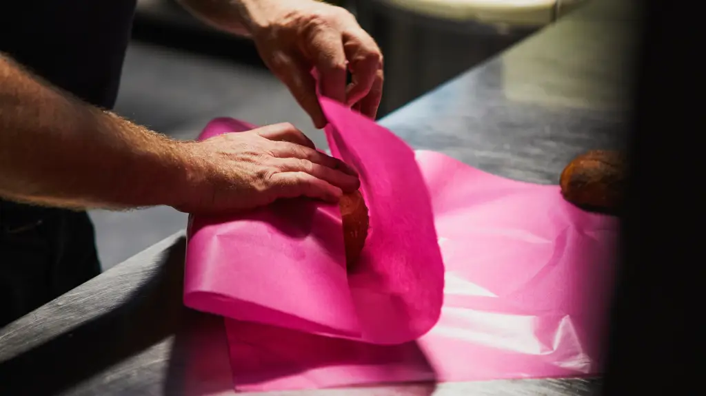 Hände verpacken ein Brot in pinkfarbenes Papier auf einer Edelstahlfläche – sorgfältiger Umgang mit dem Produkt bei bread.love