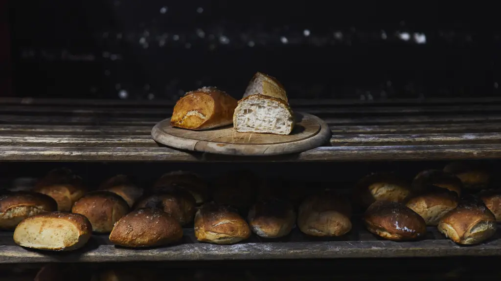 Frisch gebackene Brote auf Holzregalen, im Vordergrund ein angeschnittenes Brot mit offener Krume auf einem runden Holzbrett