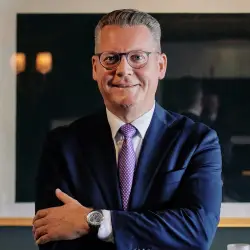 Hervé Mahler, Maître d’hôtel im Grand Hotel Les Trois Rois in Basel, im dunkelblauen Anzug mit Brille und gemusterter Krawatte vor dunklem Hintergrund